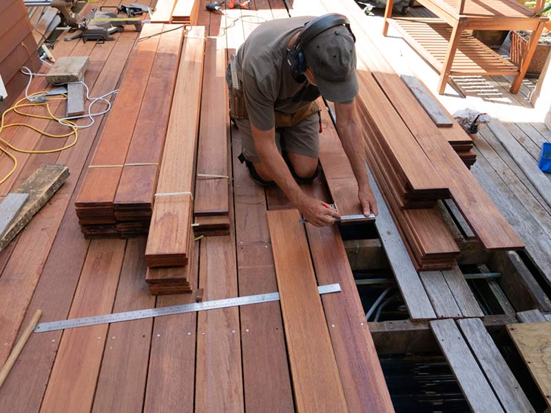 A person measures wooden planks while kneeling on a partially built deck outdoors.