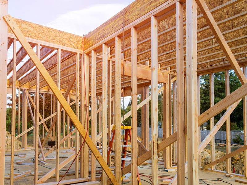 Wooden framing of a house under construction, with beams, studs, and tools visible on the ground.