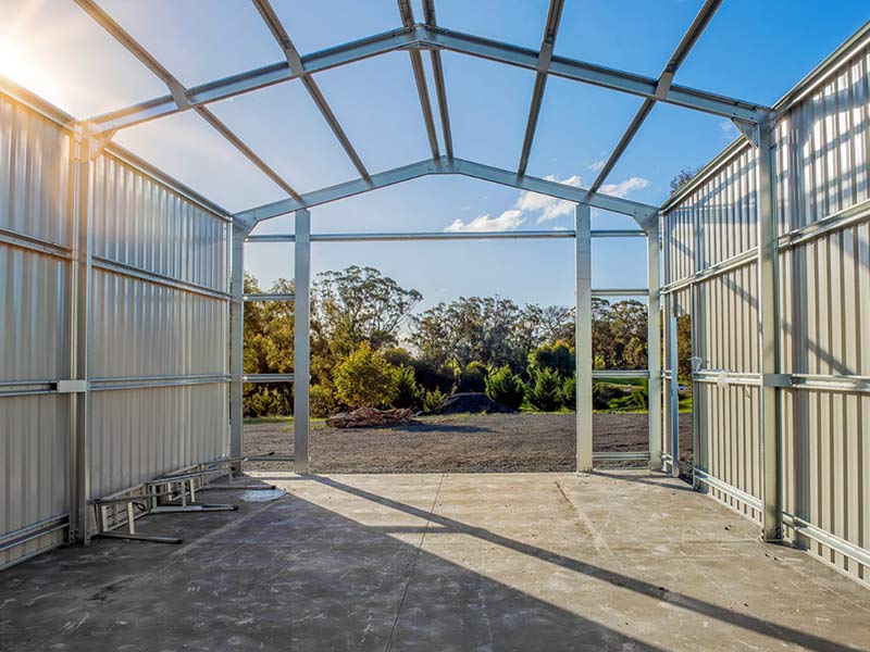 Steel-framed shed under construction with open roof, looking out to trees and blue sky.