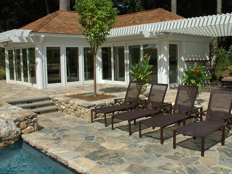 Three lounge chairs on a stone patio beside a pool, with a white pool house and pergola in the background.