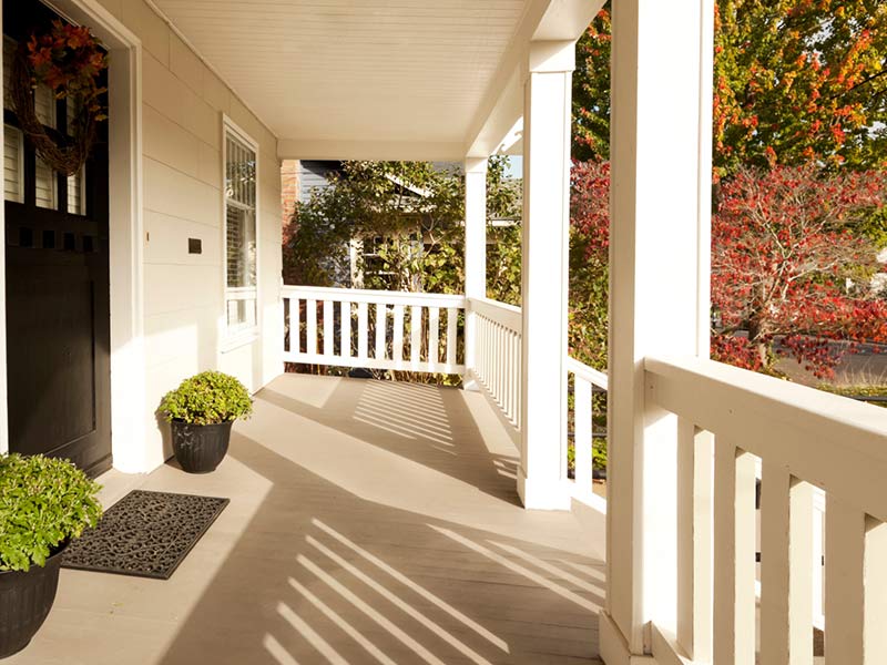 Sunny front porch with white railings, potted plants, and a view of trees with fall foliage.