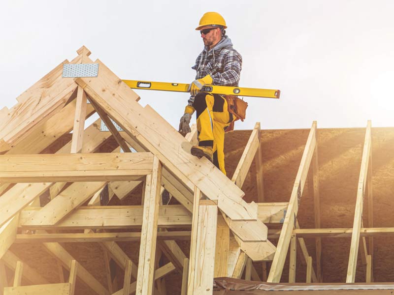 Construction worker in safety gear uses a level on the wooden frame of a roof under construction.