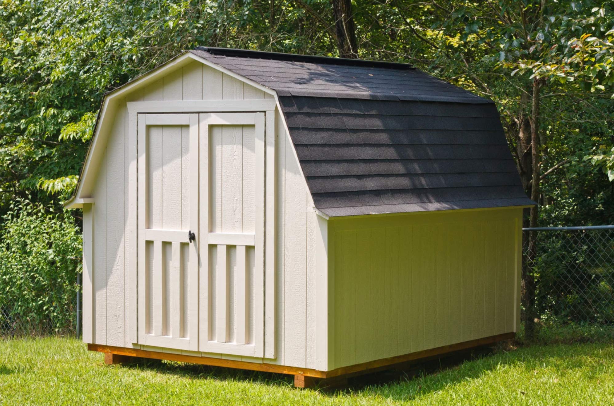 A beige wooden storage shed with double doors and a black shingle roof sits on green grass near trees.