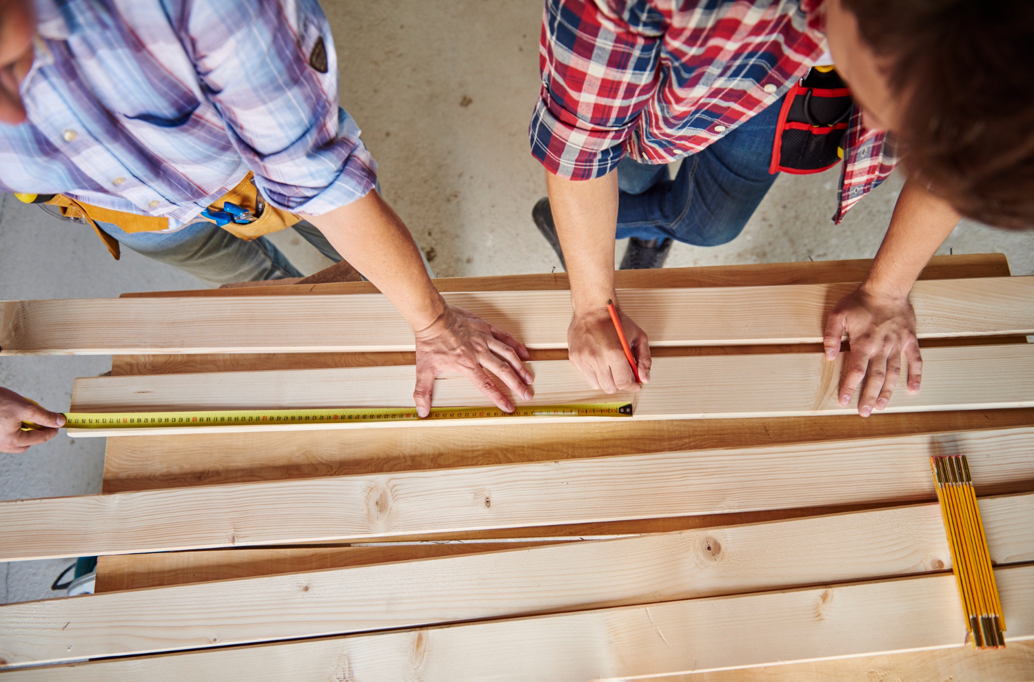 Two people measure and mark wooden planks on a workbench, preparing for a woodworking project.